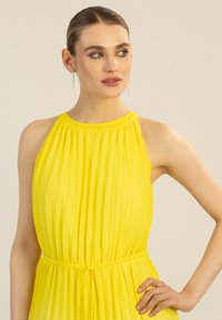 Vestido amarillo plisado con un diseño de cuello alto, tejido ligero y cintura fruncida, que presenta una textura suave y una silueta fluida.