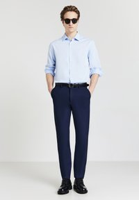 Uomo con occhiali da sole neri, camicia azzurro chiaro arrotolata, pantaloni blu navy, cintura nera e scarpe eleganti nere, in piedi con le mani nelle tasche.