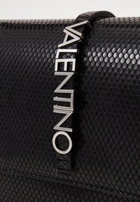 Černá texturovaná kožená kabelka s vzorem včelí plástve. Obsahuje stříbrné logo a má elegantní design. Hladké akcenty dotvářejí celkový vzhled.