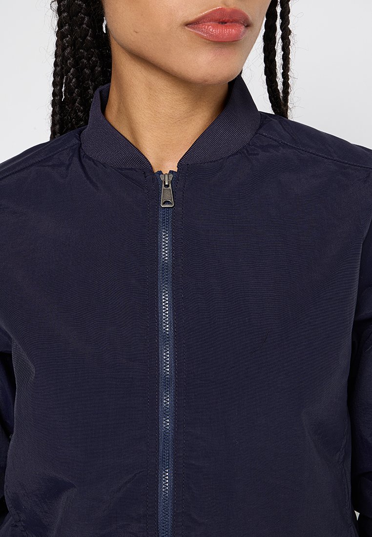 Donna con capelli intrecciati che indossa una giacca blu navy con zip e colletto a coste, fotografata dalla nuca al petto.