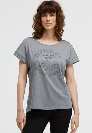 Frau mit lockigem braunem Haar, die ein graues T-Shirt mit hochgerollten Ärmeln und schwarze Hosen trägt, steht vor einem schlichten Hintergrund.