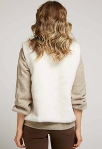 Gilet en fausse fourrure blanche avec des manches beiges. Texture douce, coutures d'épaules arrondies et ourlet côtelé. Associé à un pantalon moulant marron.