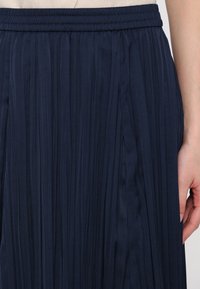 Jupe plissée bleu marine avec taille élastiquée, portée par une personne dont la main droite repose à côté de la jupe.