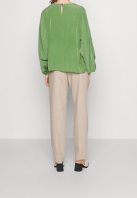 Blouse verte texturée à manches longues, avec un détail de dos à trou de serrure, assortie à un pantalon beige et une sandale à talons noirs.