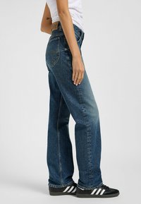 Blaue Denim-Jeans mit einer entspannten Passform, mittelhoher Taille, gelben Nähten und Gesäßtaschen. Kombiniert mit schwarzen Sneakern mit weißen Streifen.
