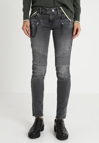 Mujer vestida con jeans gris ajustados con paneles de rodilla acanalados, detalles de cremallera y cadenas, zapatos negros de tacón grueso y un suéter verde con ribetes amarillos.