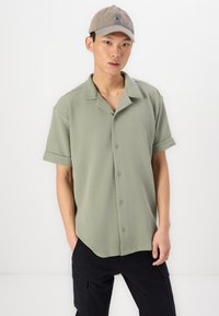 Kortärmad, button-up skjorta i ljusgrön texturerad tyg, med en camp collar och uppvikta ärmslut, tillsammans med en grå keps.