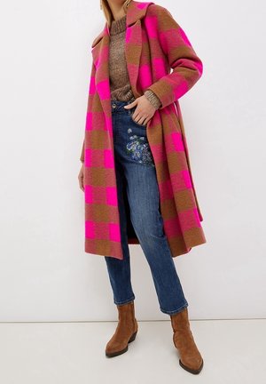 Kobieta w różowo-brązowym, długim, kratkowanym płaszczu, brązowym dzianinowym swetrze, haftowanych niebieskich dżinsach i brązowych zamszowych botkach stoi.