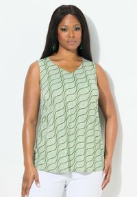 Top verde senza maniche con un motivo a onde, design a V e una vestibilità ampia. Tessuto liscio con una leggera lucentezza, abbinato a pantaloni bianchi.