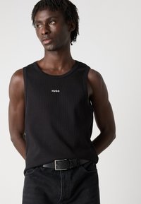 Top fără mâneci, negru, cu textură în dungi, având un logo alb "HUGO" pe partea din față. Purtați cu blugi albaștri închiși și o curea neagră.