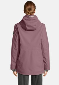 Eine Person trägt eine gedämpfte lila wasserfeste Jacke mit Kapuze und einem Aufnäher an den Ärmeln, gesehen von hinten vor einem schlichten Hintergrund.