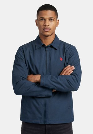 Junger Mann mit kurzen Haaren, der eine marineblaue Jacke mit Reißverschluss und rotem Logo trägt, steht mit verschränkten Armen vor weißem Hintergrund.