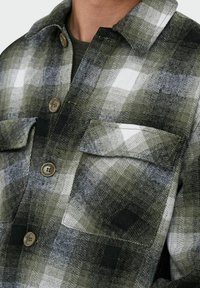 Camisa a cuadros con botones, que presenta colores verde, blanco y negro, hecha de un tejido texturizado, con dos bolsillos en el pecho y botones marrones.
