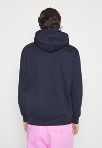 Persona con cabello oscuro y ondulado, vistiendo una sudadera azul marino y pantalones rosa claro, de espaldas contra un fondo claro y liso.