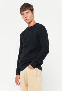 Giovane uomo che indossa un maglione a maglia nera e pantaloni beige, con una mano in tasca, su uno sfondo bianco uniforme.