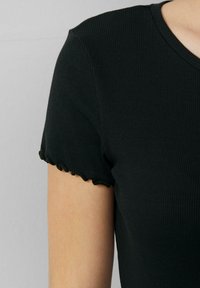 Nahaufnahme einer Person, die ein schwarzes, geripptes Kurzarmoberteil mit Rüschenärmeln und Rundhalsausschnitt vor einem schlichten Hintergrund trägt.