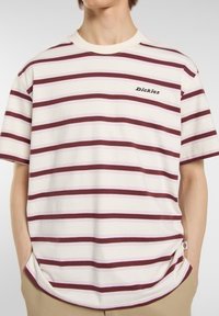 Model in a striped t-shirt featuring maroon and pink stripes, with the "Dickies" logo displayed on the chest. Hands in pockets.
