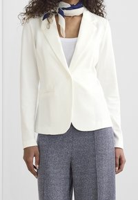 Bílý blazer s klopami, jedním knoflíkem a předními kapsami. Nosený přes světlejší top a vzorované kalhoty.