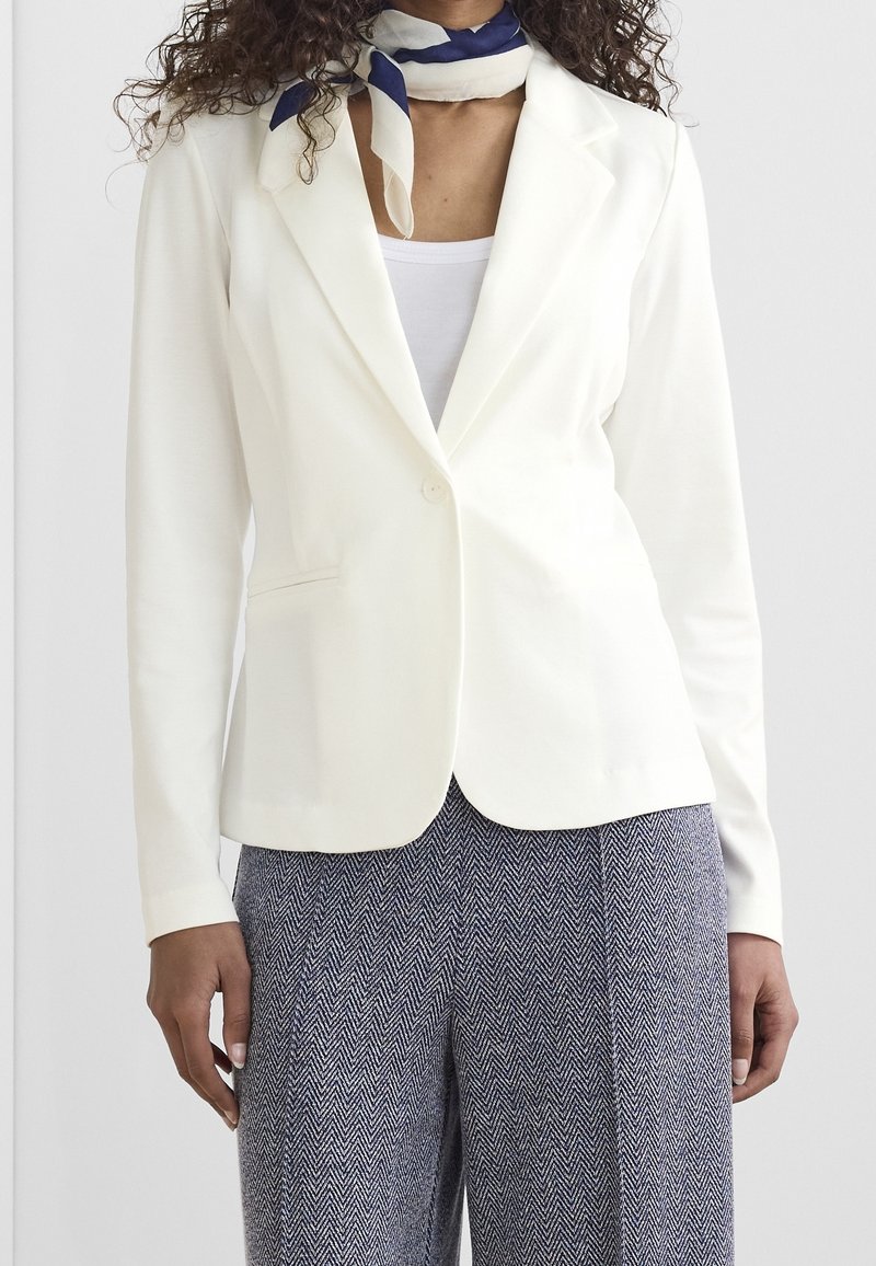 Bílý blazer s klopami, jedním knoflíkem a předními kapsami. Nosený přes světlejší top a vzorované kalhoty.