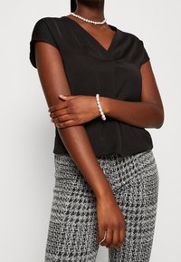 Top nero a maniche corte con scollo a V, abbinato a pantaloni grigi a fantasia e a un bracciale di perle, che mette in evidenza un tessuto liscio e una vestibilità slim.