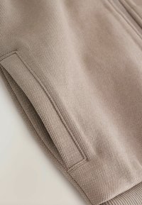 Pantalons en tissu marron clair avec un tissage texturé, comportant des poches latérales et des poignets côtelés. Le matériau semble doux et durable.