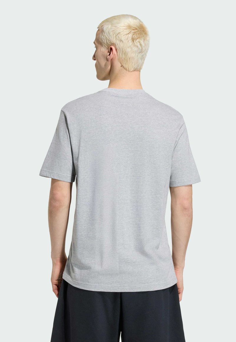 Lyshåret person set bagfra iført en ensfarvet lysegrå kortærmet T-shirt og løse sorte shorts mod en ensfarvet lys baggrund.