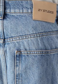 Jeans en denim bleu clair avec une texture lisse, des coutures orange contrastantes et une étiquette en cuir beige portant la mention "2Y STUDIOS" sur la taille.