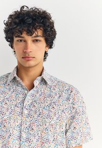 Chemise à manches courtes à fond blanc, ornée de motifs colorés de fleurs et de feuilles dans des tons de violet, orange et vert.