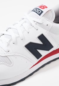 Tenisky New Balance 500 s bílým koženým a síťovaným svrškem, námořnicky modrým logem, červenými akcenty a bílými tkaničkami na texturované podrážce.