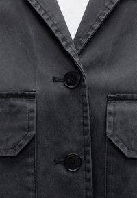 Gros plan sur le devant d'une veste gris foncé avec deux boutons noirs, col à revers cranté et deux poches à rabat.
