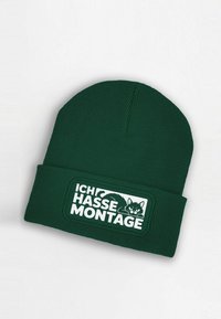 Green ribbed knit beanie featuring a white rectangular patch with text "ICH HASSE MONTAGE" and a black cat graphic.