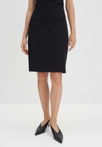 Schwarzer knielanger Bleistiftrock mit glatter Textur, hoher Taille und seitlichen Taschen, kombiniert mit spitzen schwarzen Schuhen.