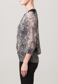 Przezroczysta czarna i biała bluzka z wzorem gałęzi drzew, okrągłym dekoltem i teksturowanym elastycznym paskiem w talii. Długie, luźne rękawy.