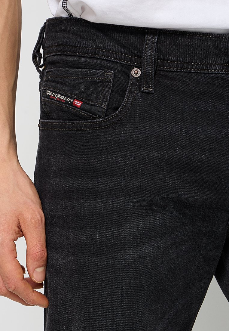 Nahaufnahme einer Person, die schwarze Diesel-Jeans trägt, mit sichtbarem Fronttaschen- und Markenetikett, die Hand ruht neben dem Oberschenkel.