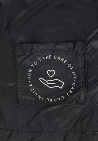 Schwarzes Stoffpatch mit einem weißen Icon einer Hand, die ein Herz hält, umgeben von kreisförmigem Text: "Wie man sich um mich kümmert, Pflegeetikett innen."