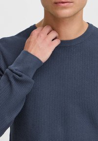 Tamsiai mėlynas tekstūruotas megztinis su apvaliu iškirpimu ir banguotomis rankogaliais. Medžiaga turi tinklelio raštą, suteikiantį gylį ir matmenį.