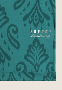Tessuto teal con un motivo damascato in tonalità di teal più scure. Include il testo "FREDDY training" in argento metallico. Texture morbida, design casual.