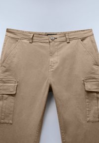 Beige cargo-bukser lavet af bomuld, med to sidelommer, lynlås i bukserne og knaplukning. Glat tekstur med syede detaljer.