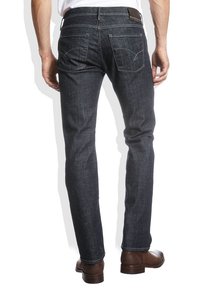 Hombre con jeans ajustados de color azul oscuro y botas de cuero marrón, visto desde atrás sobre un fondo blanco.