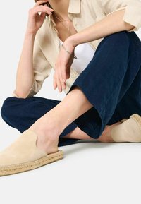 Kobieta w beżowych espadrylach na płaskiej podeszwie, granatowych szerokich spodniach, beżowej koszuli z guzikami i srebrnej biżuterii, siedząca na podłodze.