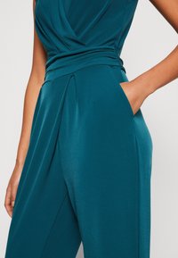 Mujer vistiendo un mono satinado color teal con cintura plisada y bolsillo lateral, con una mano apoyada dentro del bolsillo.