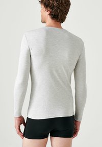 Camicia a maniche lunghe grigia chiara realizzata in un tessuto morbido, con un design aderente, scollo rotondo e orlo pulito, indossata sopra pantaloni corti neri.