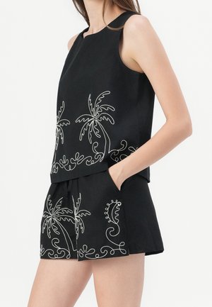 Woman wearing black sleeveless top and shorts with white embroidered palm tree and swirl patterns, hand in shorts pocket.