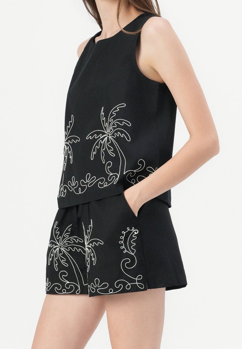 Femme portant un haut noir sans manches et un short avec des motifs brodés blancs de palmier et de spirale, main dans la poche du short.