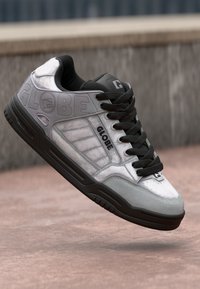 Skate-style sneaker in gray and black with "GLOBE" branding on side, black laces, and textured sole, suspended above a paved surface.