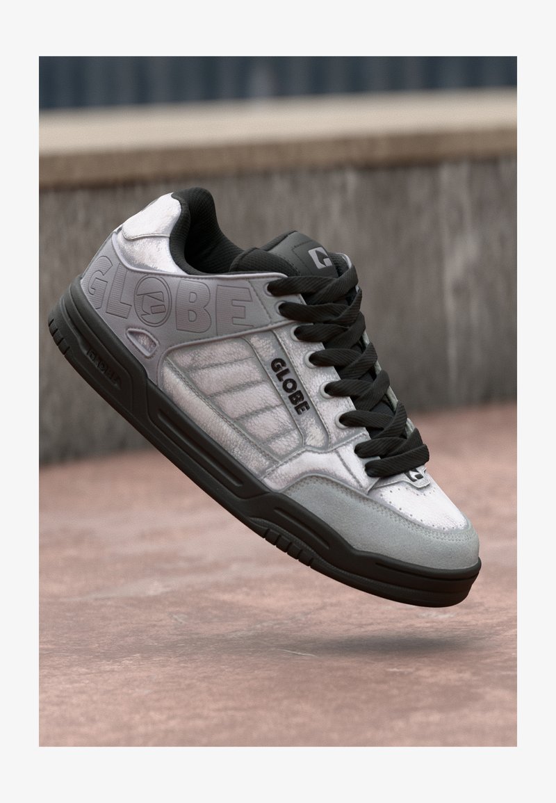 Sneaker in stile skate in grigio e nero con il marchio "GLOBE" sul lato, lacci neri e suola testurizzata, sospesa sopra una superficie pavimentata.