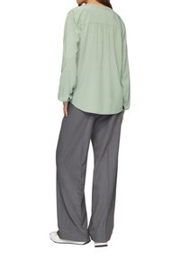 Chemise à manches longues vert clair avec détail froncé au dos, associée à un pantalon gris ample et des baskets blanches, présentant une coupe décontractée.