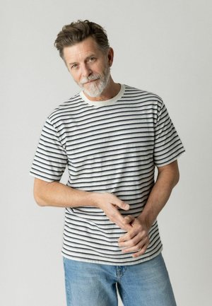 Middelaldrende mand med gråt skæg og bølget hår iført en hvid og sort stribet T-shirt og blå jeans, stående mod en ensartet lys baggrund.