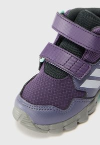 Lila Sport Schuh mit einer strukturierten Synthetik-Oberfläche, schwarzen Mesh-Akzenten, zwei Klettverschlüssen und einer durchsichtigen grauen Gummisohle.