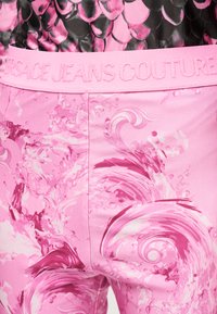 Ροζ παντελόνι με στροβιλιζόμενα αφηρημένα μοτίβα νερού και ζωνάρι μέσης με ανάγλυφη την επιγραφή "Versace Jeans Couture" σε ρόδινη κλωστή που ταιριάζει.